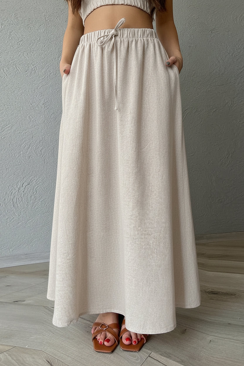 A person wearing a long, loose-fitting maxi skirt in a light beige or oatmeal linen-like fabric, paired with a matching ribbed crop top and brown sandals, standing against a textured gray wall and light wood floor.